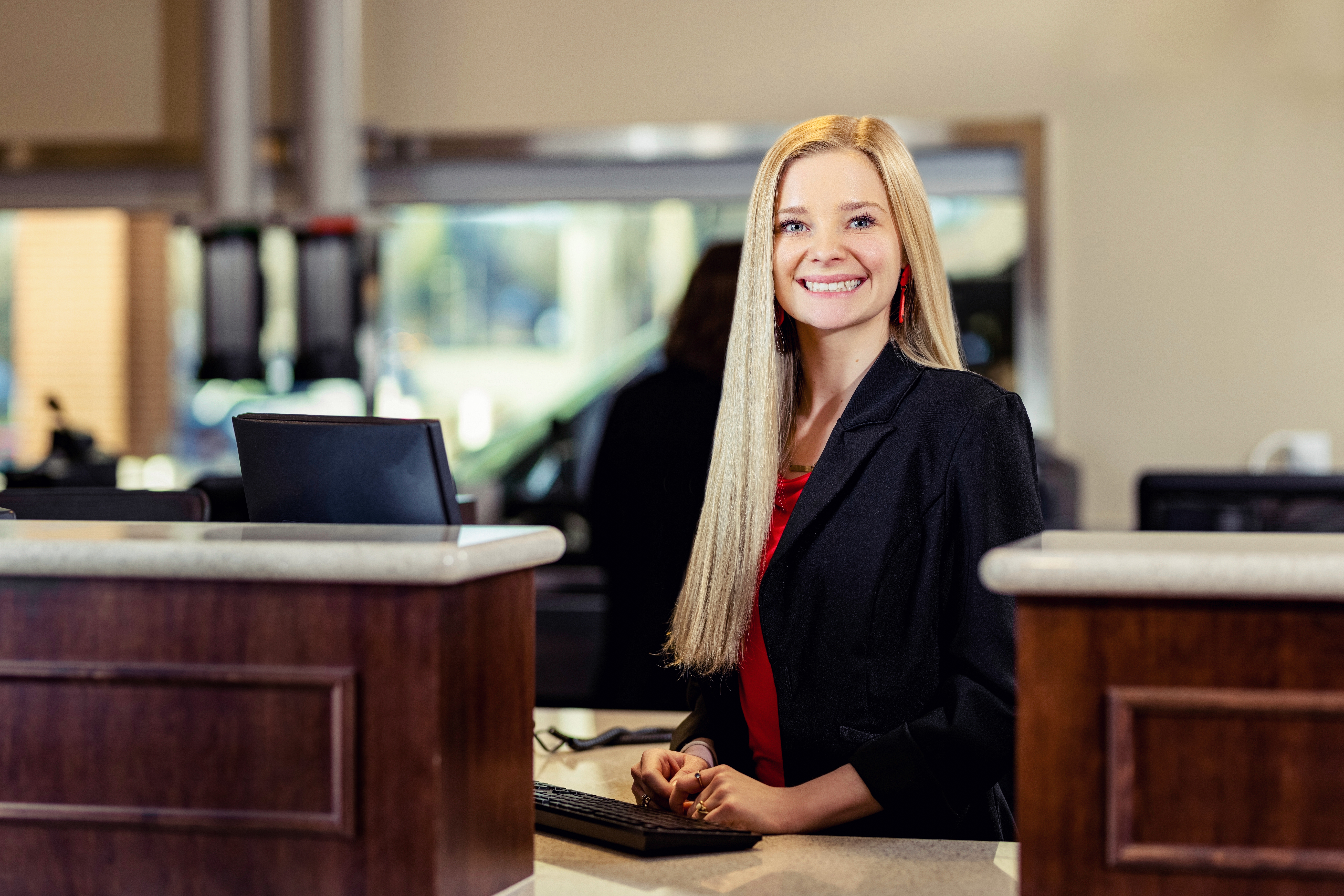 Blonde Woman in red shirt and black blazer smiling behind a computer