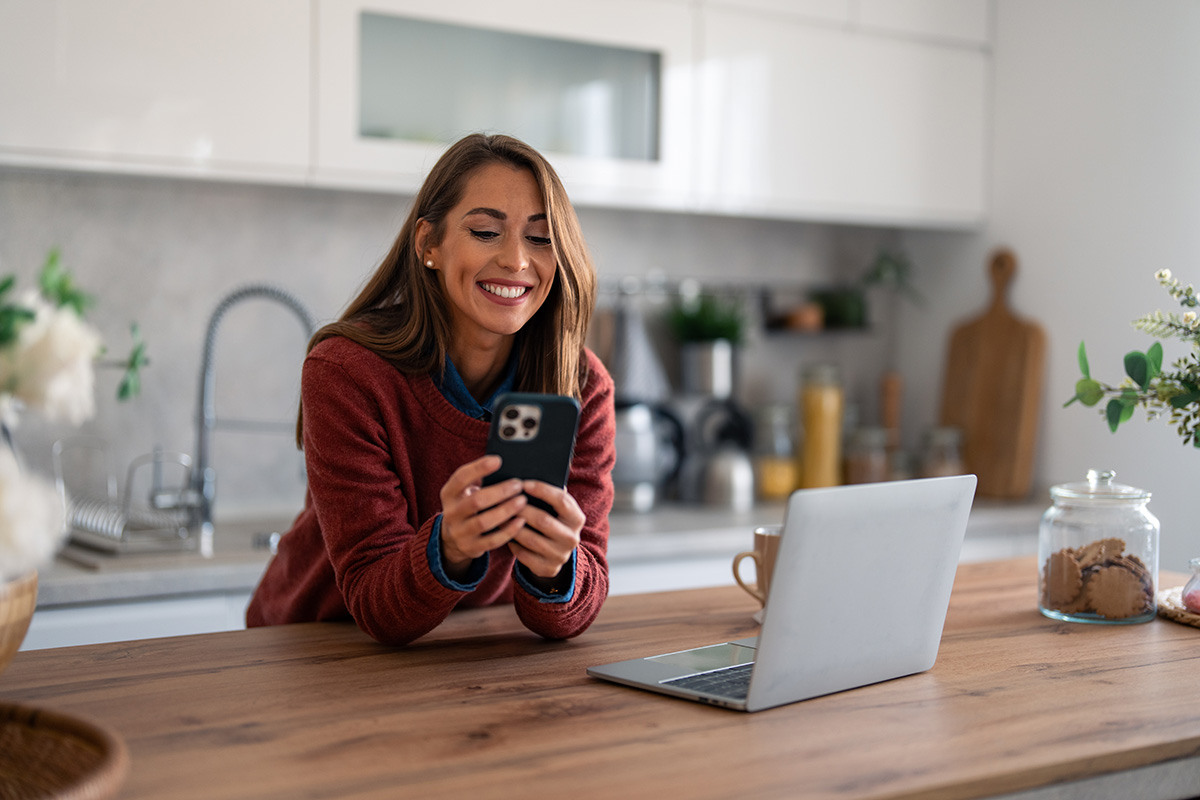 A woman smiles while engaging with her phone and laptop, showcasing a moment of joy in her digital interaction.